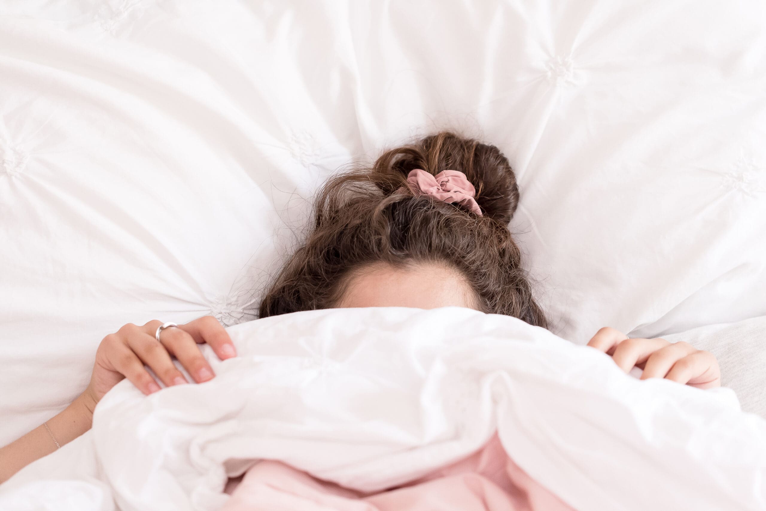 Woman with messy bun and pink scrunchie peeking out from under white bedding, holding the duvet up over her face in a cozy bedroom setting -Laura Keenan-Nervous System Coach